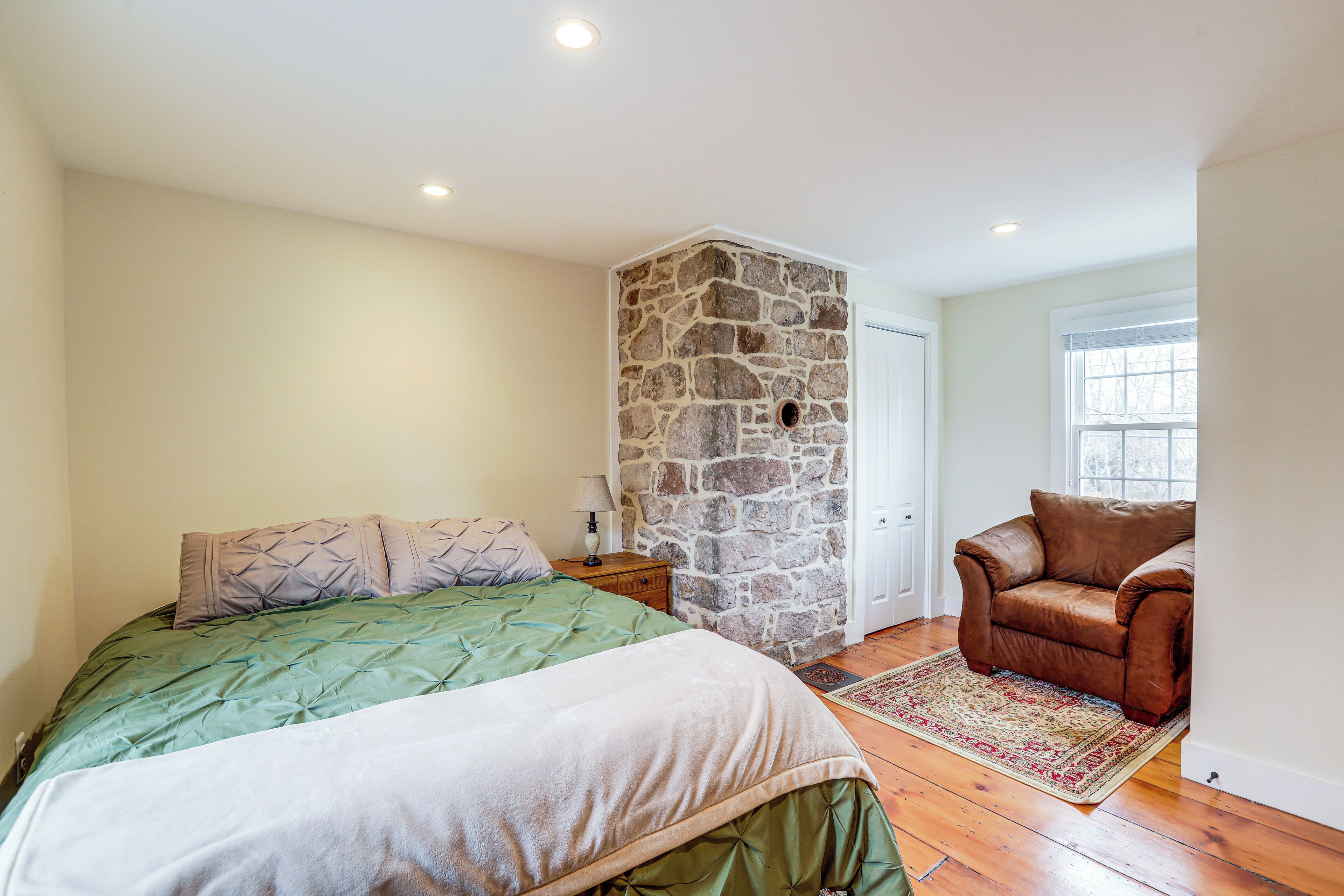 A bedroom with a bed, a stone wall, a brown leather chair, and a window.