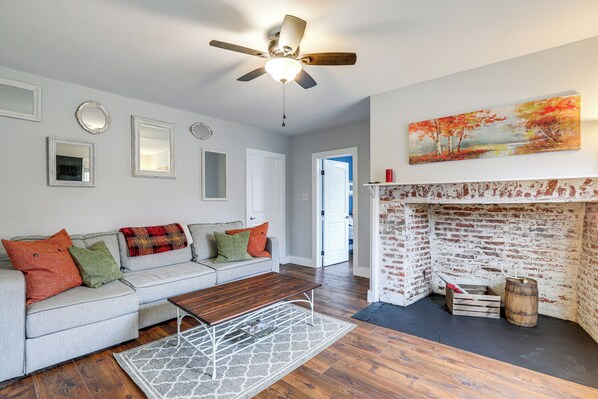 A living room with a gray sofa, wooden coffee table, and a fireplace with a brick wall. - In-town & Walkable: Historic Yardley Retreat! (Morrisville)
