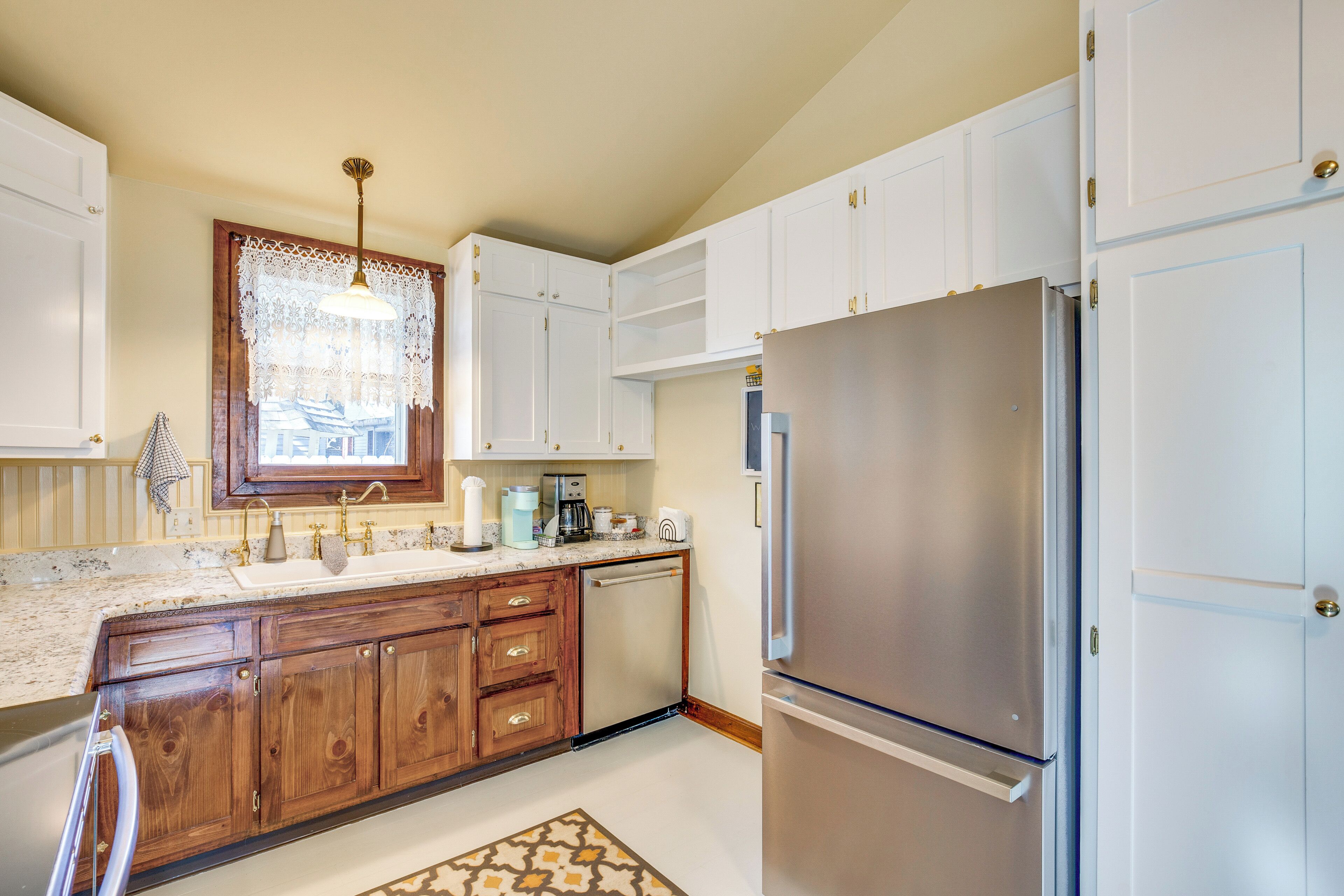 A kitchen with white cabinets, a stainless steel refrigerator, a dishwasher, and a window with a lace curtain.