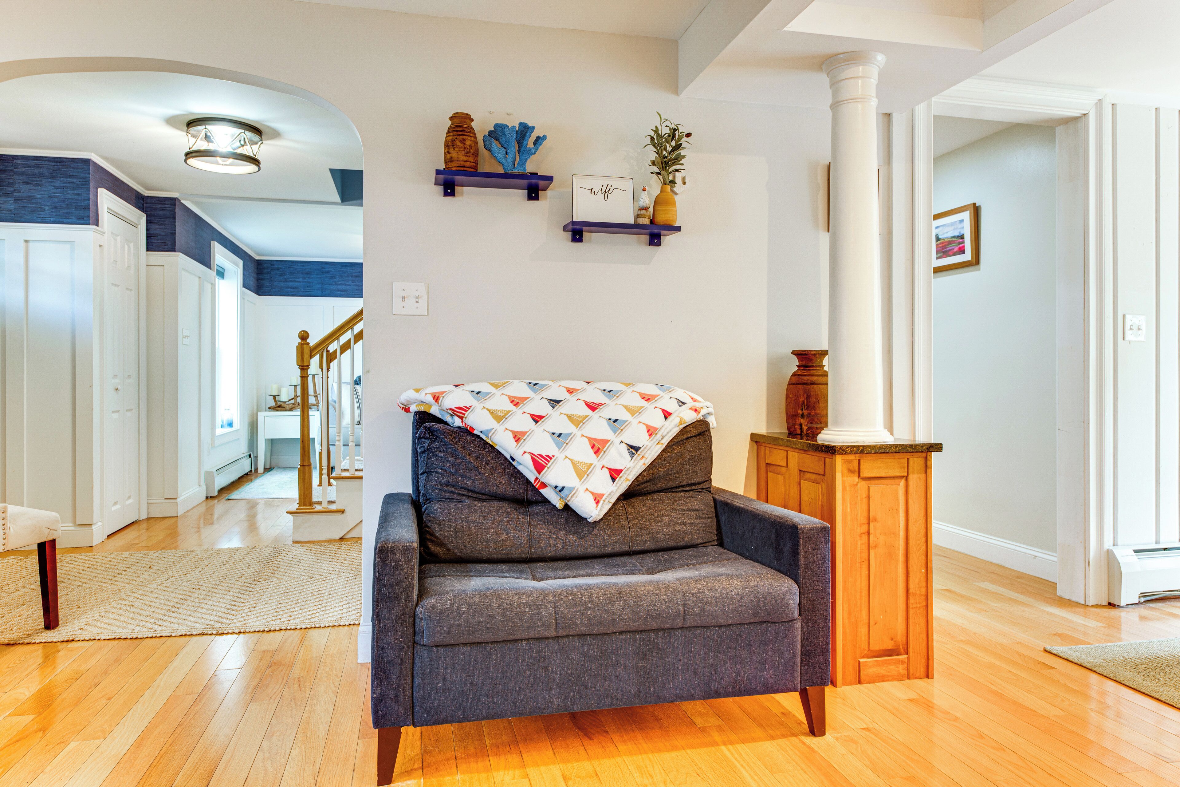 A living room with a grey sofa, wooden flooring, a white pillar, and a staircase leading to another room.