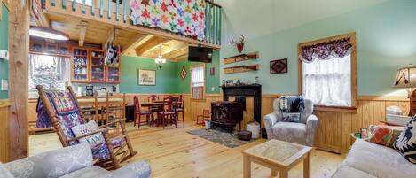 Un acogedor interior de cabaña con piso de madera, chimenea, comedor y un altillo con colcha de colores.