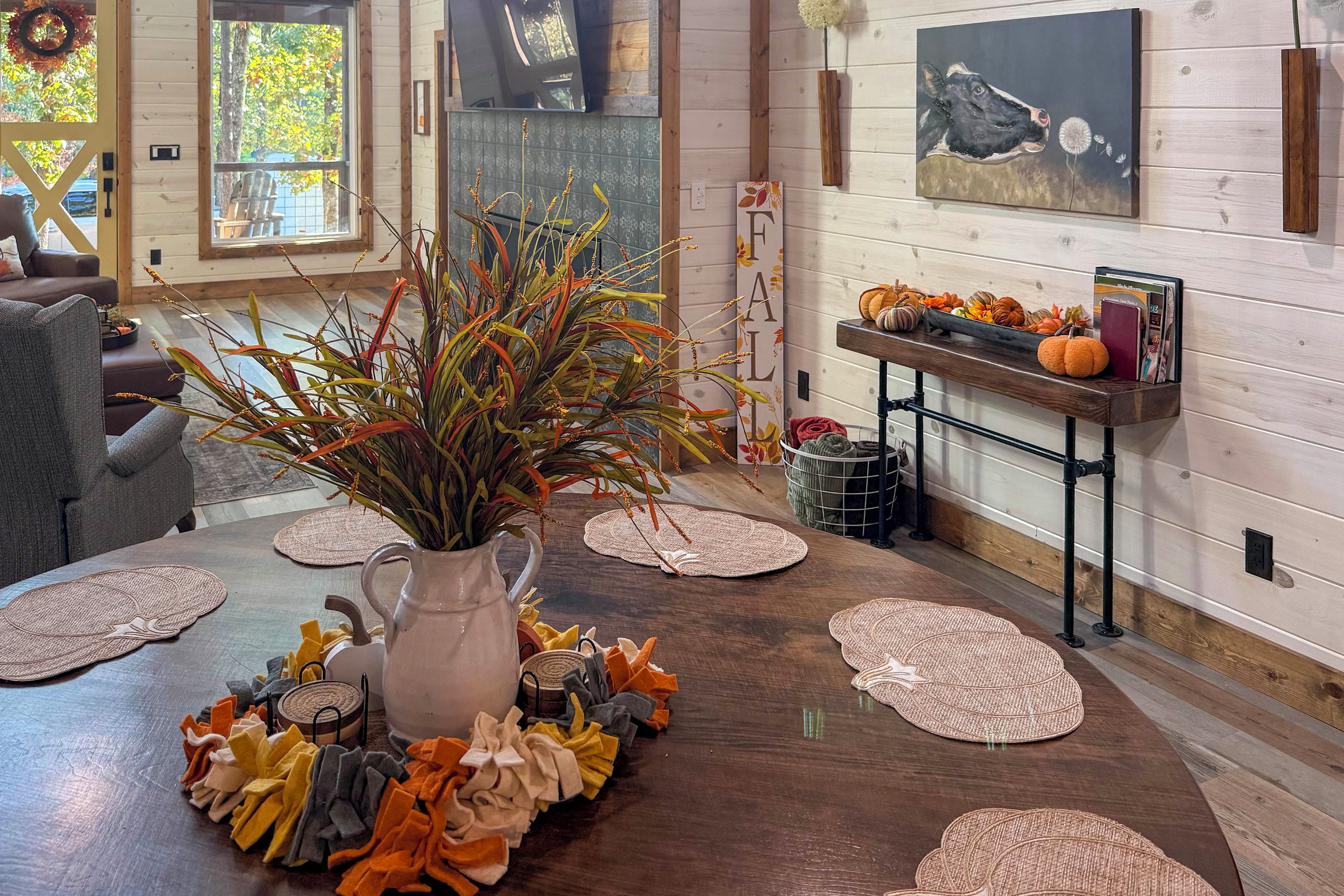 A dining area with a wooden table, a vase with autumn leaves, and a wall with a painting and a sign.