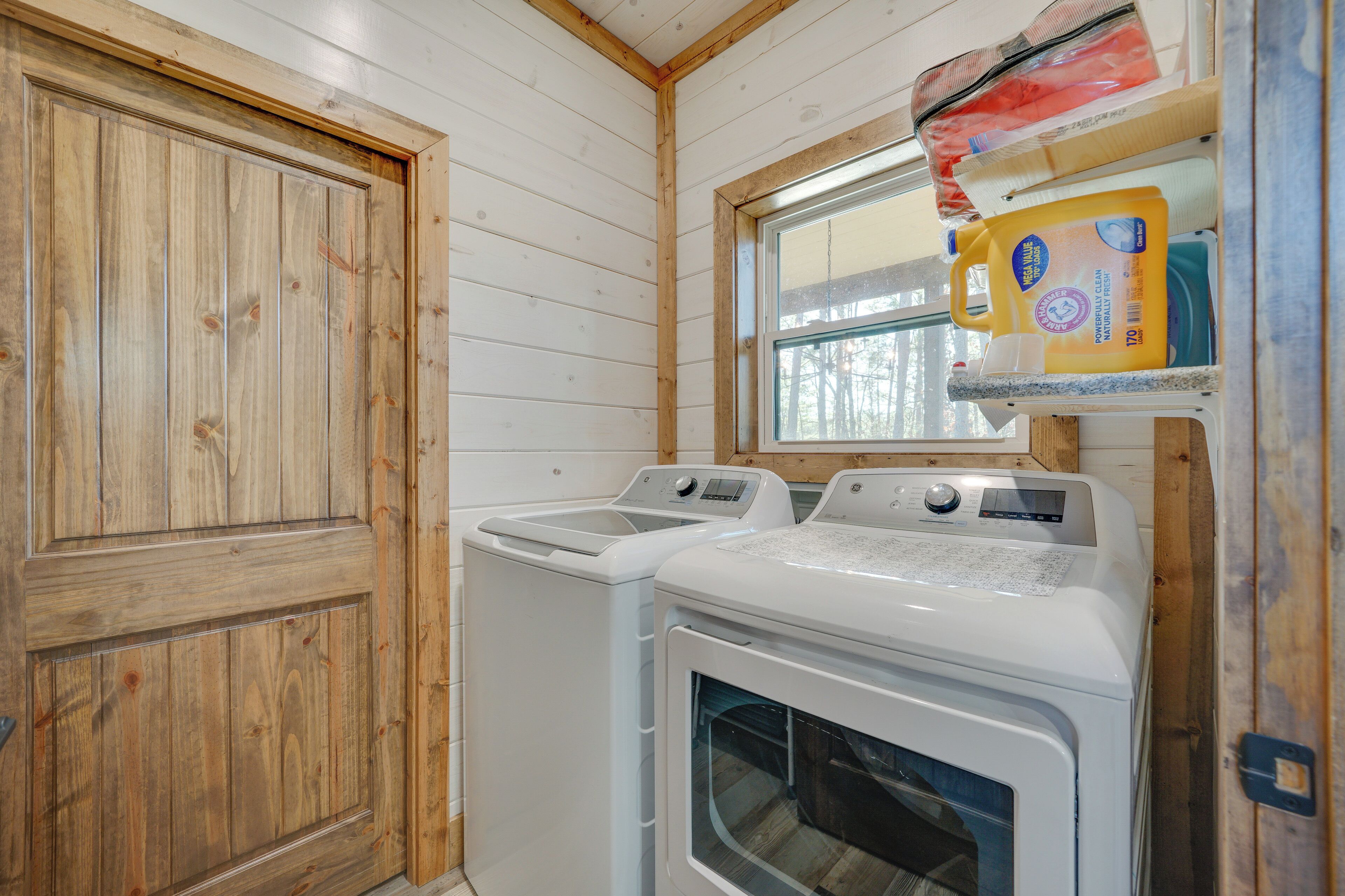 A laundry room with two washing machines and a dryer, wooden walls, and a window with a view of trees.