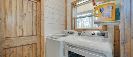 A laundry room with two washing machines and a dryer, wooden walls, and a window with a view of trees.