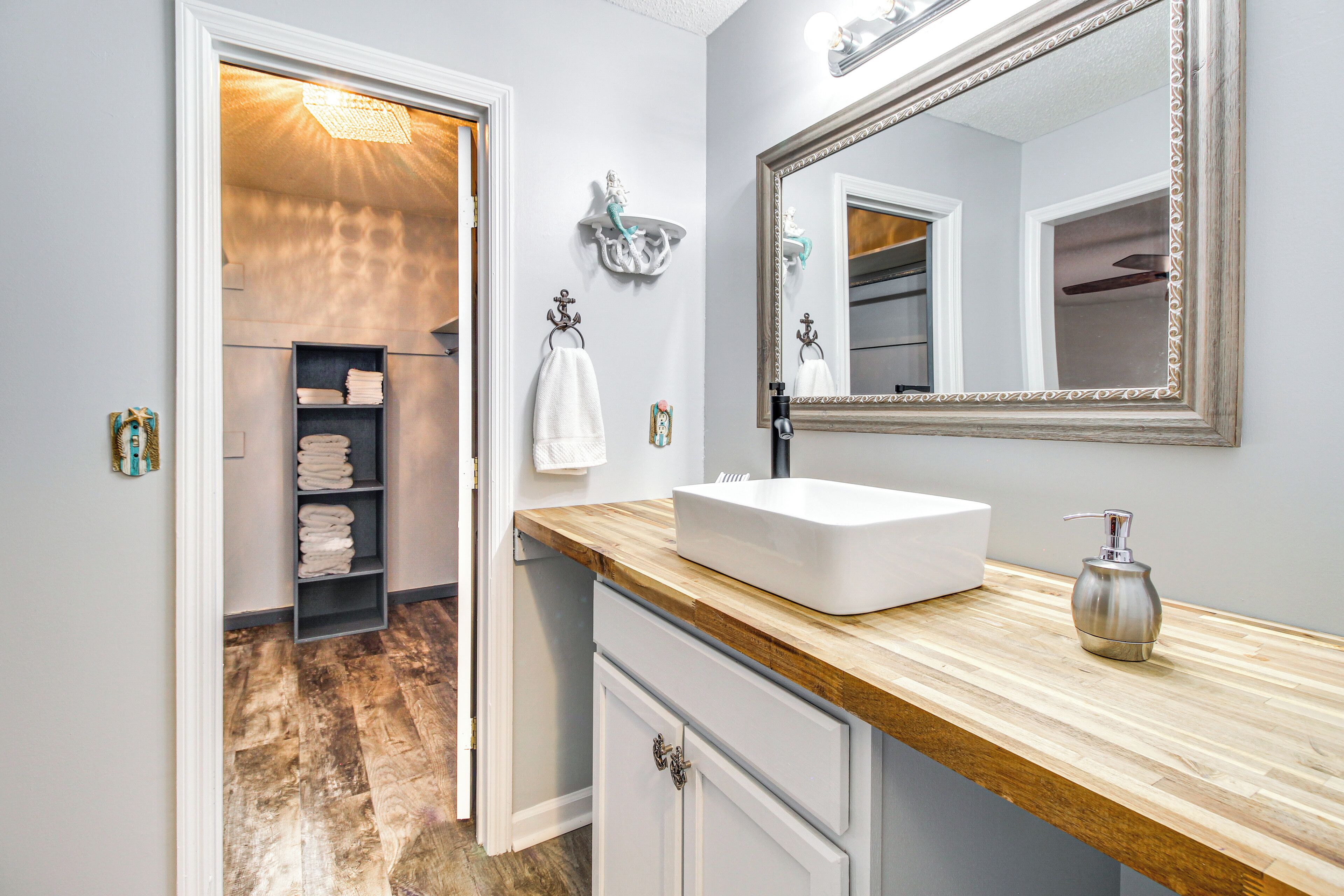 A bathroom with a wooden vanity, white sink, and a large mirror. There is a towel rack with folded towels and a shelf with more towels.