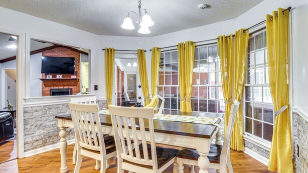 A dining area with a brick fireplace, a TV mounted on the wall, and yellow curtains on the windows. - Home 2.5 Mi to Fort Stewart w/ Heated Pool (Hinesville)