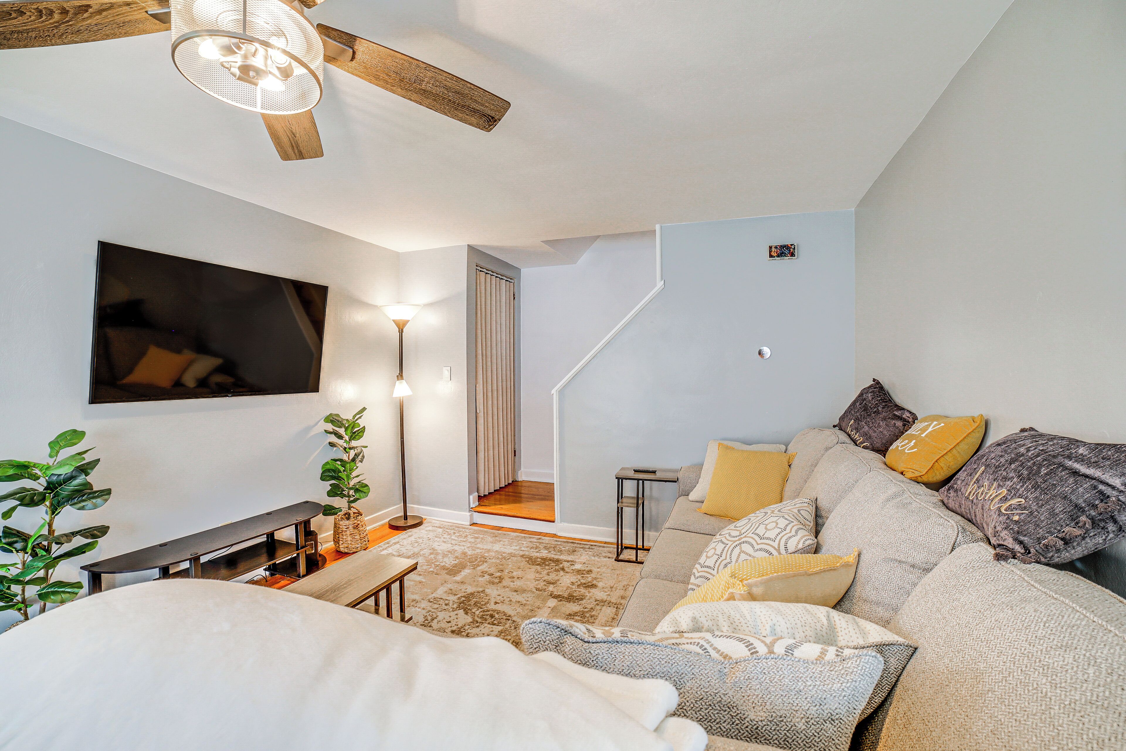 A modern living room with a sofa, a TV mounted on the wall, a ceiling fan, and a staircase leading to another level.