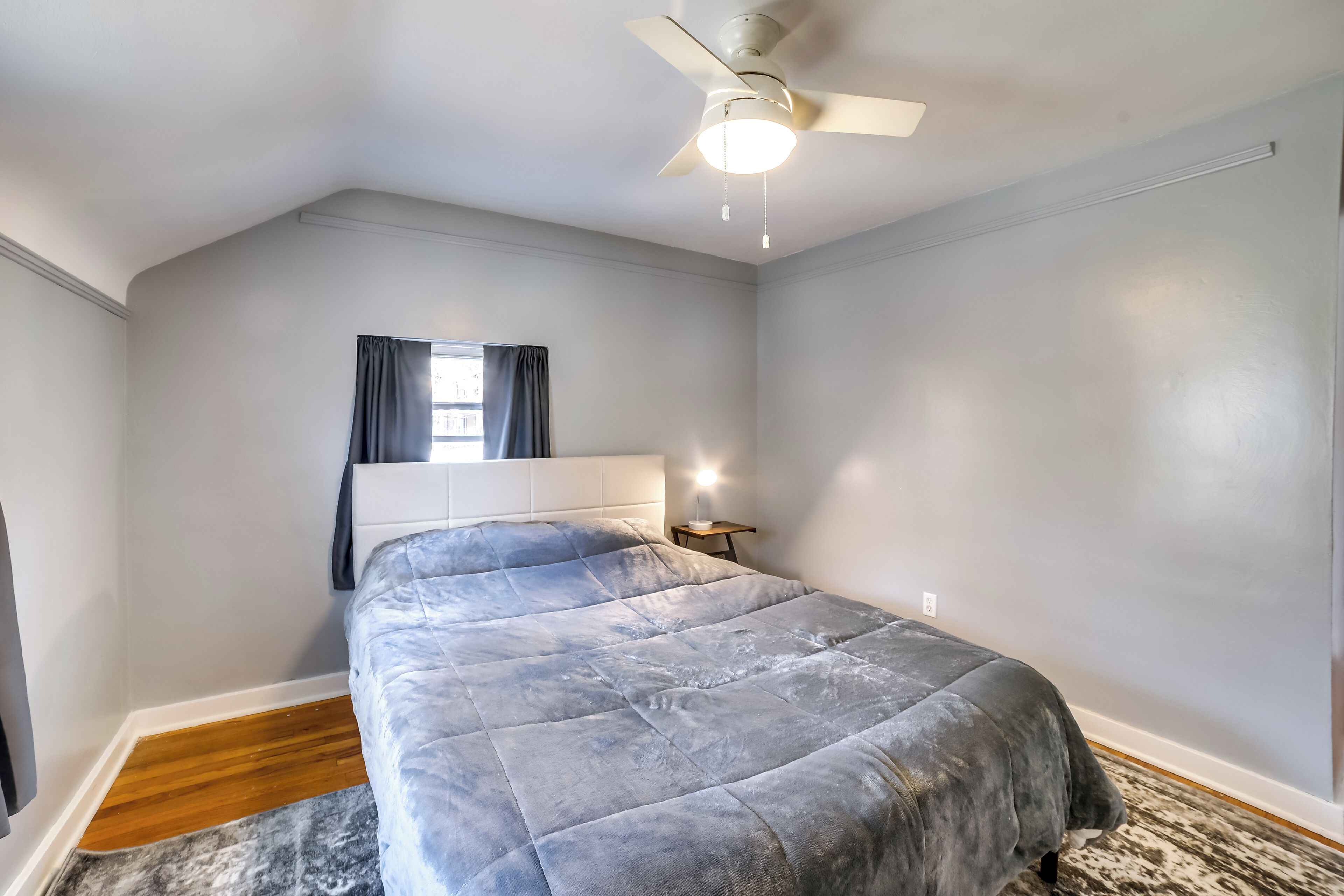 Un dormitorio con cama, ventilador de techo, mesita de noche y ventana con cortinas.