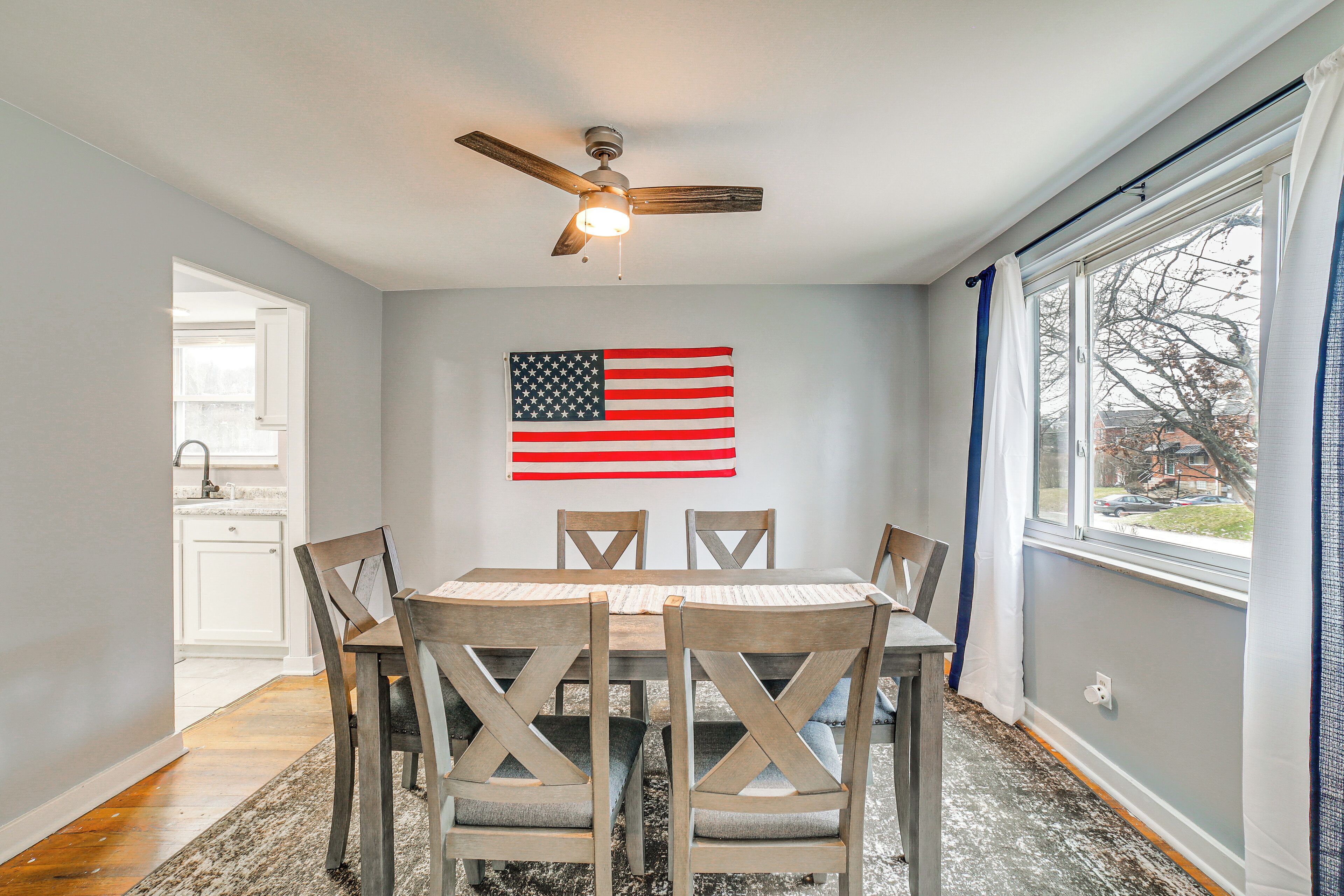 Un coin repas avec une table et des chaises, un ventilateur de plafond, une fenêtre avec des rideaux et un drapeau américain accroché au mur.