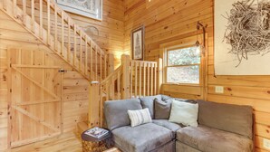 A wooden cabin interior with a gray sofa, a wooden staircase, and a framed picture of a pine tree. - 'toccoa Waterfront Hideaway:' Hot Tub & Fire Pit! (Mineral Bluff)
