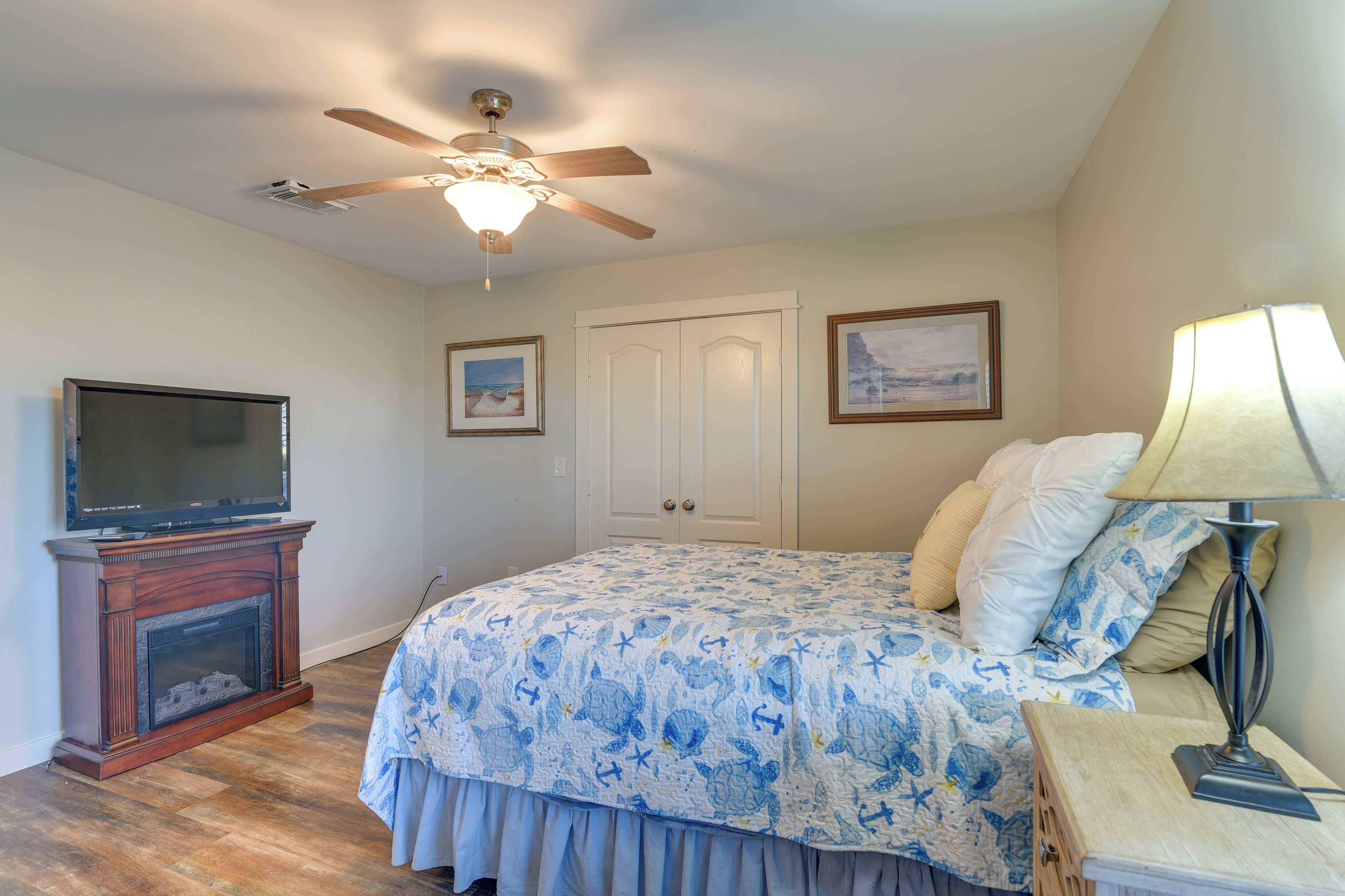 A bedroom with a bed, a television, a ceiling fan, a fireplace, and framed pictures on the wall.