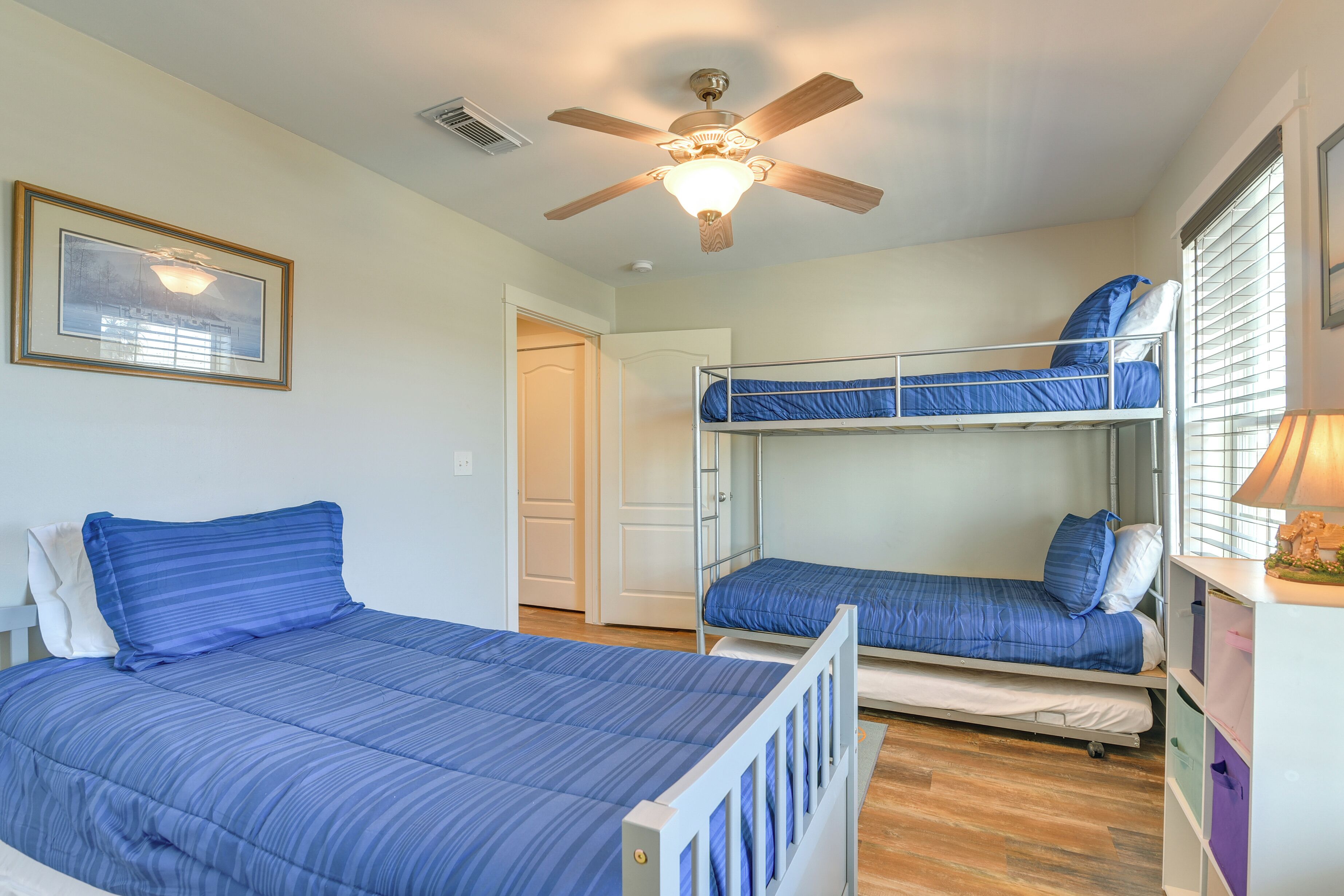 A room with two bunk beds, a ceiling fan, a framed picture, and a window with blinds.