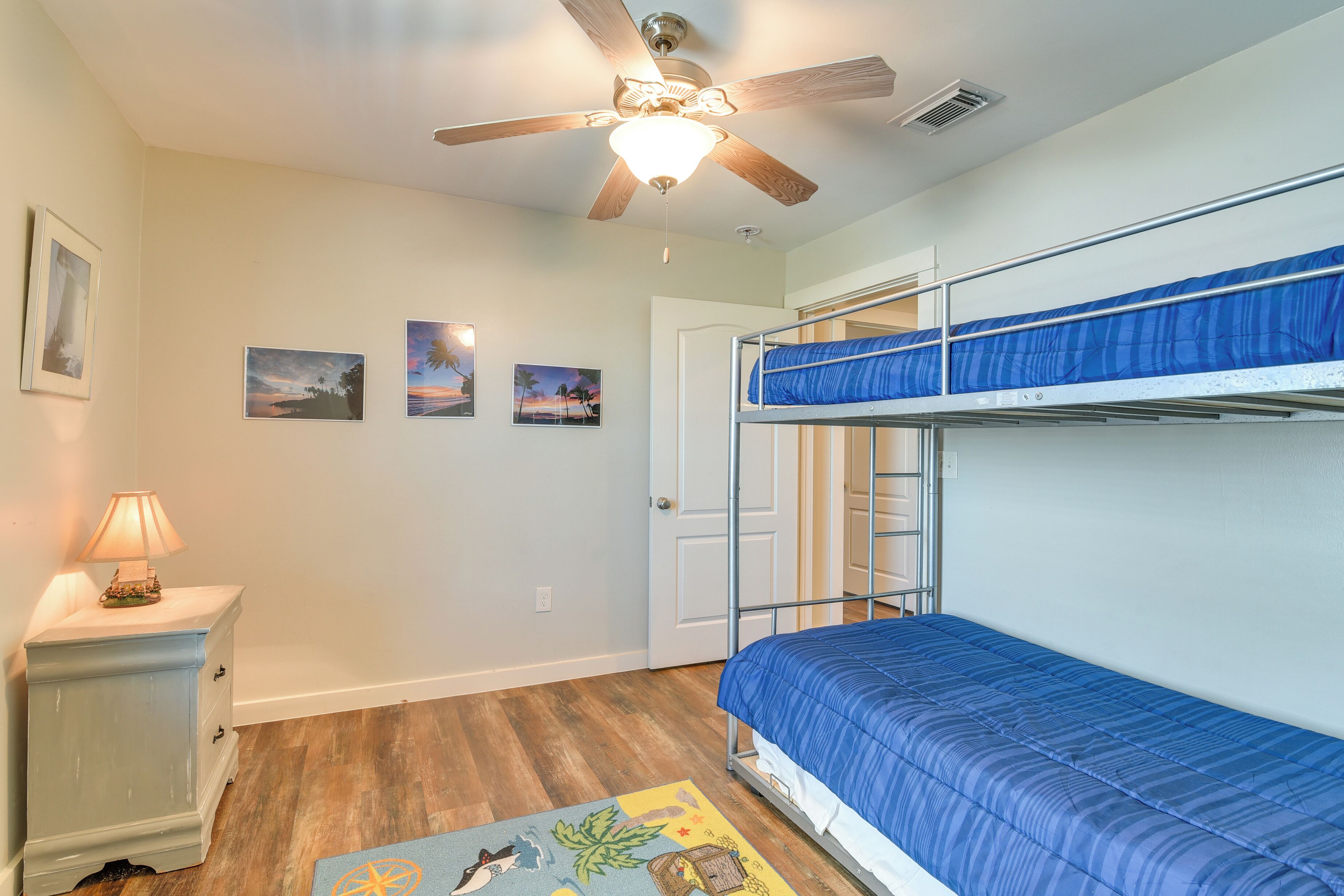 A room with bunk beds, a ceiling fan, a nightstand with a lamp, and framed pictures on the wall.