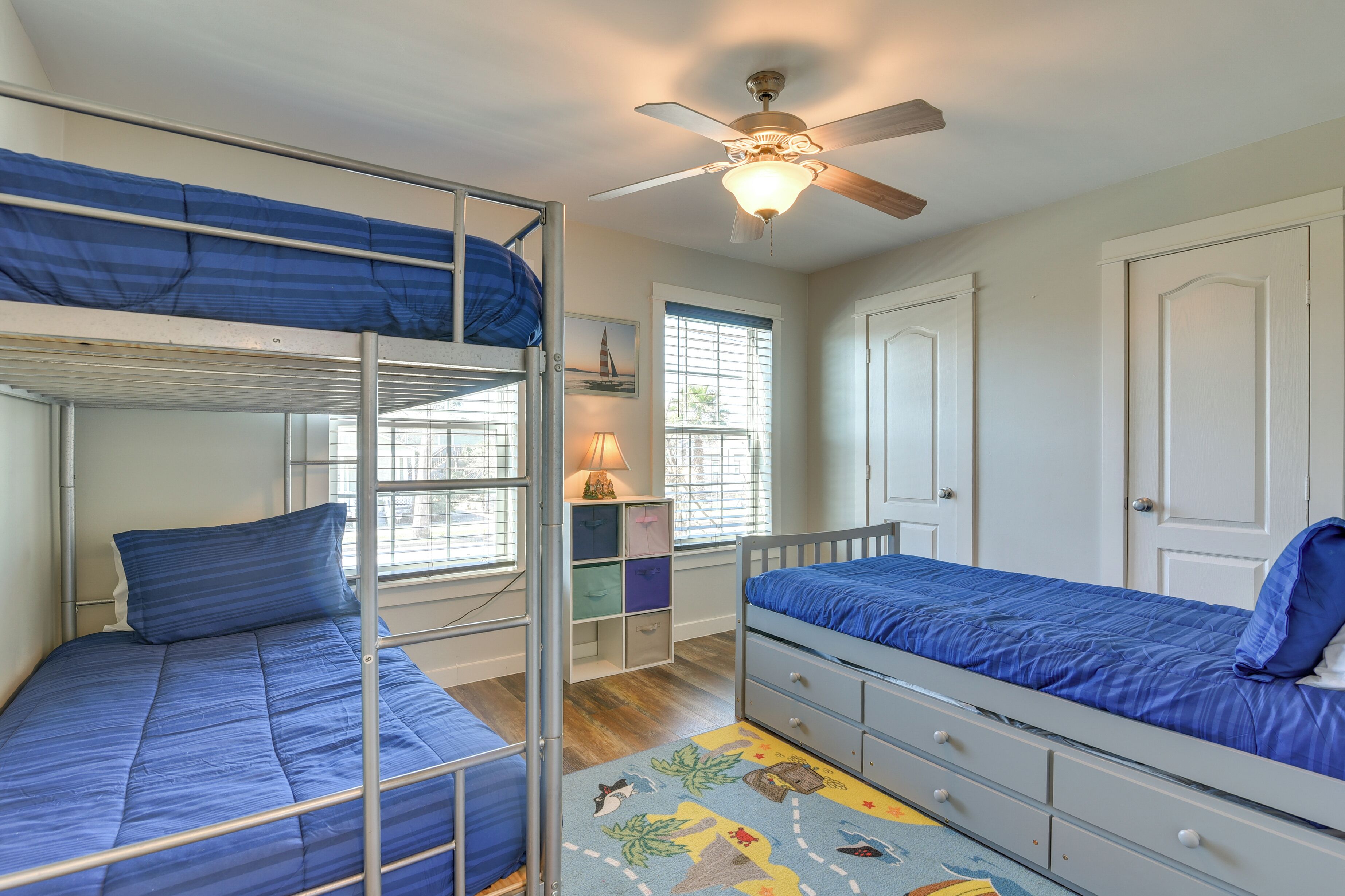 A bunk bed room with a ceiling fan, a window with blinds, a desk with drawers, and a door.