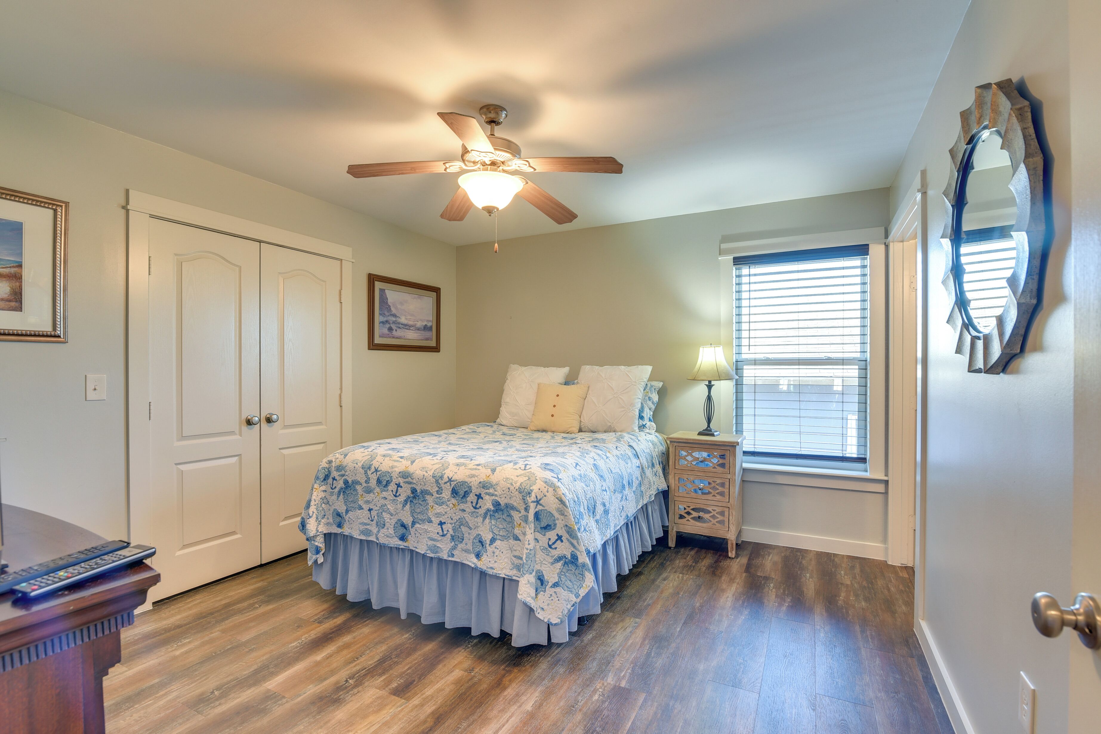 A bedroom with a bed, a ceiling fan, a nightstand, a lamp, a window with blinds, and a door with a mirror.