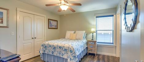 A bedroom with a bed, a ceiling fan, a nightstand, a lamp, a window with blinds, and a door with a mirror.