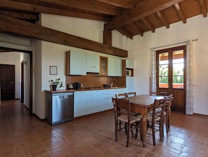 Cozinha com vigas de madeira, mesa de jantar com cadeiras, máquina de lavar louça e janela com cortinas.