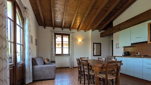 A cozy room with a wooden ceiling, a dining table with chairs, a sofa, a window with curtains, and a kitchen area. - Villa Santa Caterina - Apartment, terrace overlooking the garden + swimming pool (Manerba del Garda)