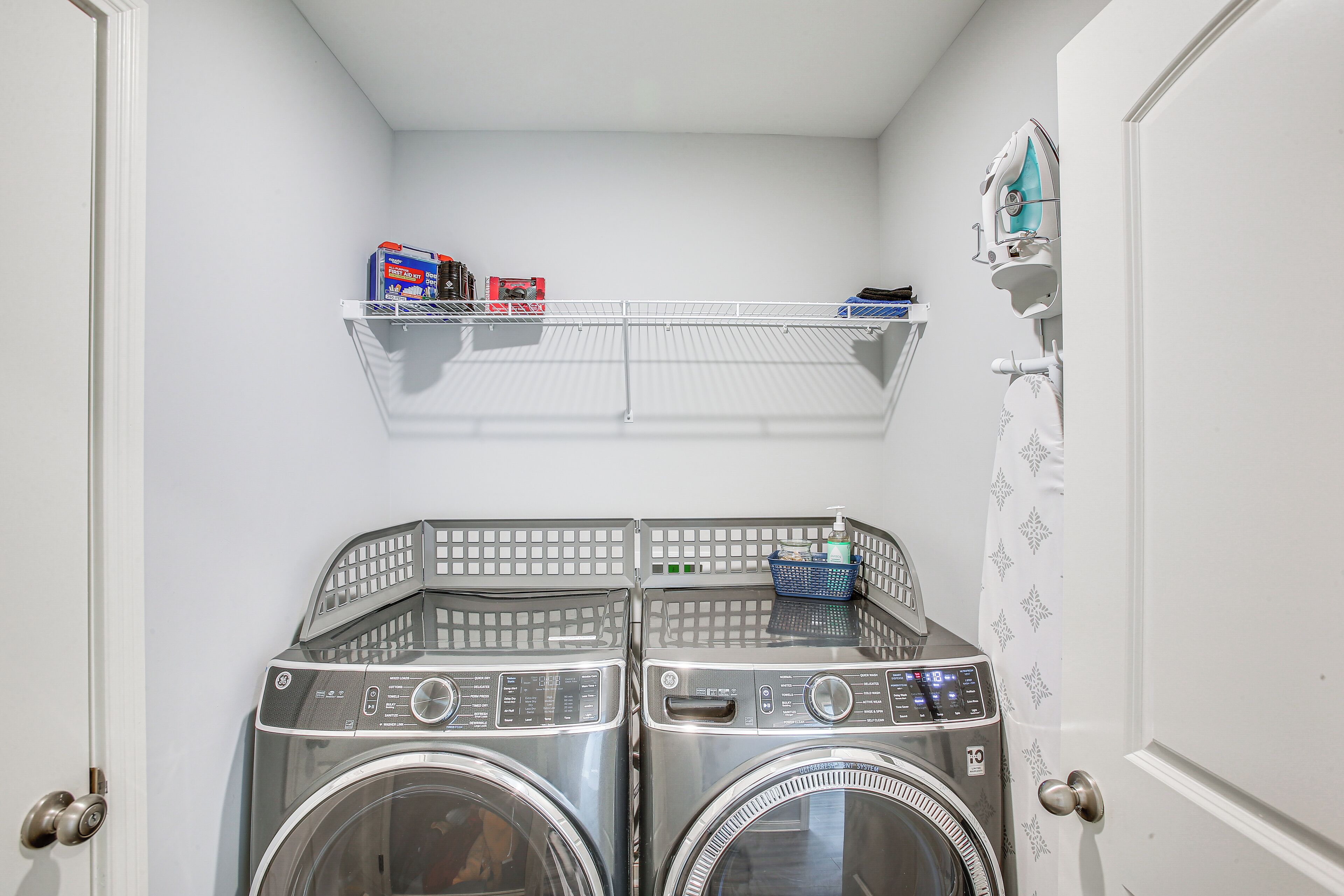 Une buanderie équipée de deux machines à laver et de deux sèche-linge à chargement frontal superposés, d’une étagère en métal contenant des produits de lessive et d’une porte ornée d’un motif décoratif.