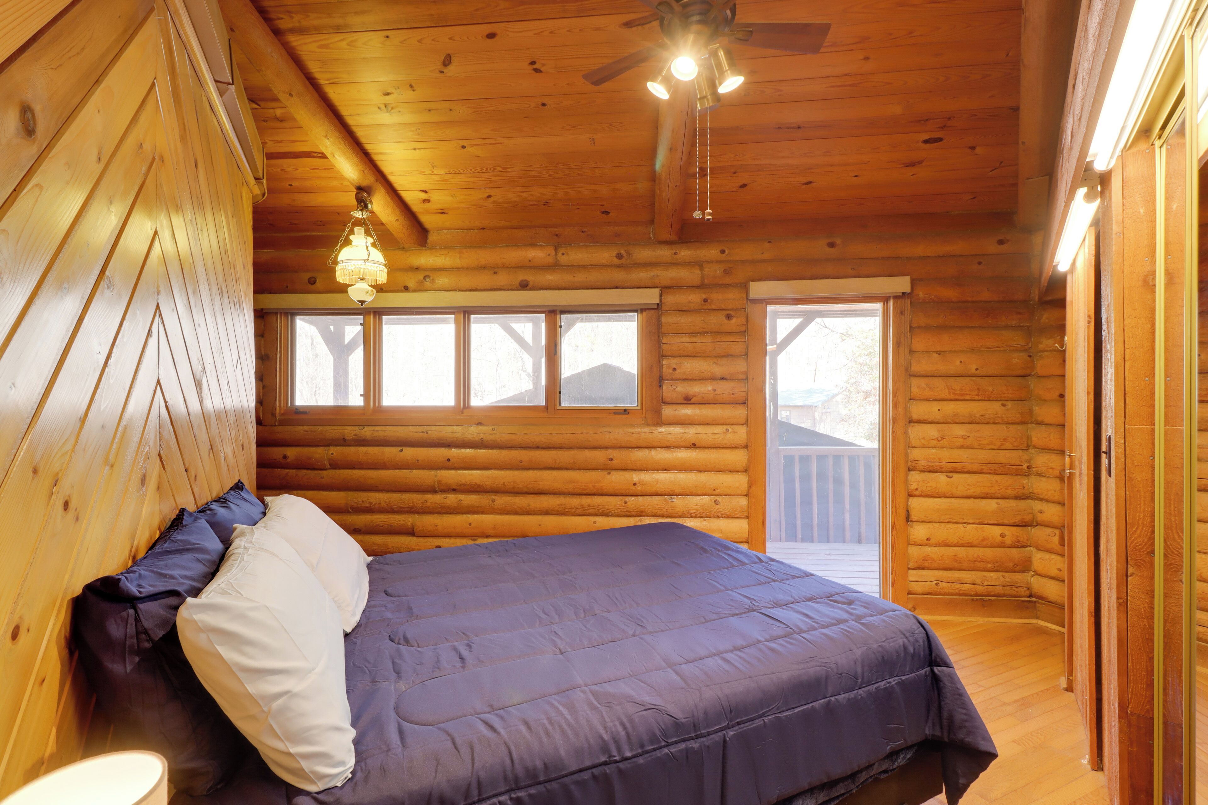 Un dormitorio de cabaña de madera con una cama, un ventilador de techo y ventanas grandes.