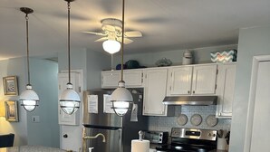 Une cuisine dotée de plans de travail en granit, de meubles blancs, d’un réfrigérateur en acier inoxydable, d’une cuisinière et d’un ventilateur de plafond. On y trouve trois tabourets de bar roses et des luminaires suspendus au-dessus du plan de travail.