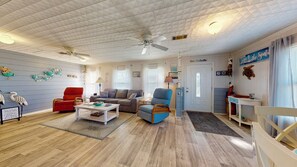 Uma sala de estar com piso de madeira, um ventilador de teto, uma cadeira vermelha, uma poltrona azul, um sofá cinzento e uma mesa de centro branca.