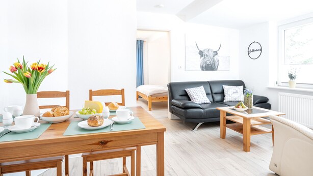 A bright, modern living room with a wooden dining table set for breakfast, a black leather sofa, and a large window with a view. - Vacation in the countryside - adventure and relaxation - what (Schellhorn)