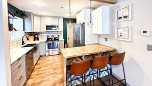 A modern kitchen with white cabinets, stainless steel appliances, a wooden island with bar stools, and pendant lighting. - Wheelhouse: Lakefront 2BD/3BA/Deck/Pool/Pet Friendly-6PP (Lake Ozark)