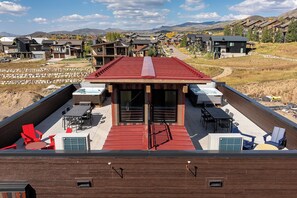 En moderne udendørs terrasse med rødt tag, flere siddeområder og udsigt til et boligområde med huse og bjerge i baggrunden.