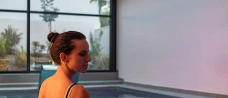 A woman sitting by an indoor pool with a large window in the background.
