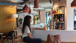 Una persona seduta al bancone di un bar, con un drink, circondata da mobili in legno e luci a sospensione.