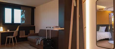 A modern hotel room with a wooden floor, a desk with a chair, a sofa, a bed, and a large mirror.