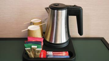 Una tetera eléctrica de acero inoxidable sobre una bandeja negra con mantel verde, que contiene varias cápsulas de café y un sobre rojo.