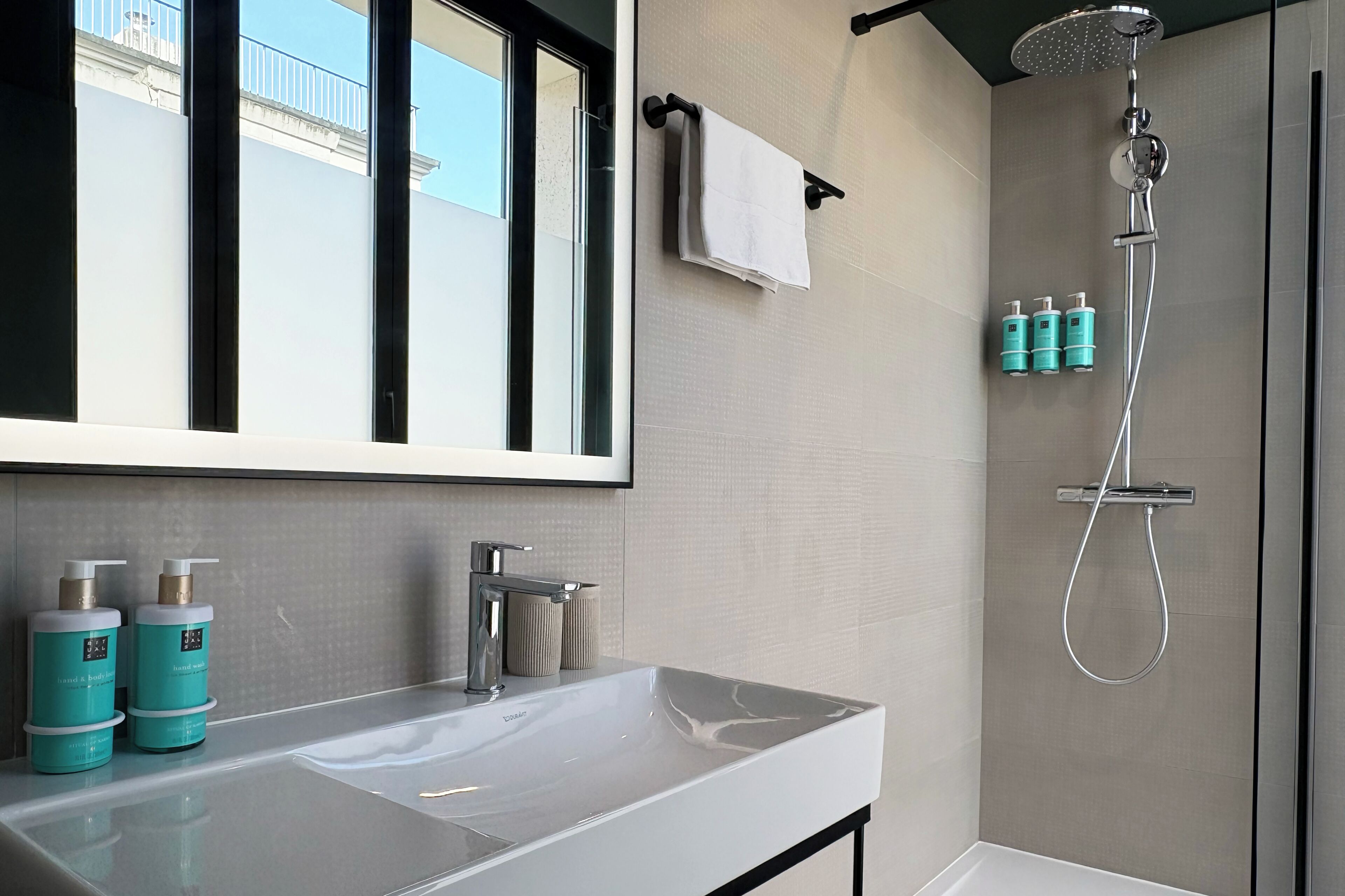 A modern bathroom with a large mirror, a white sink, a shower, and two teal bottles on the counter.