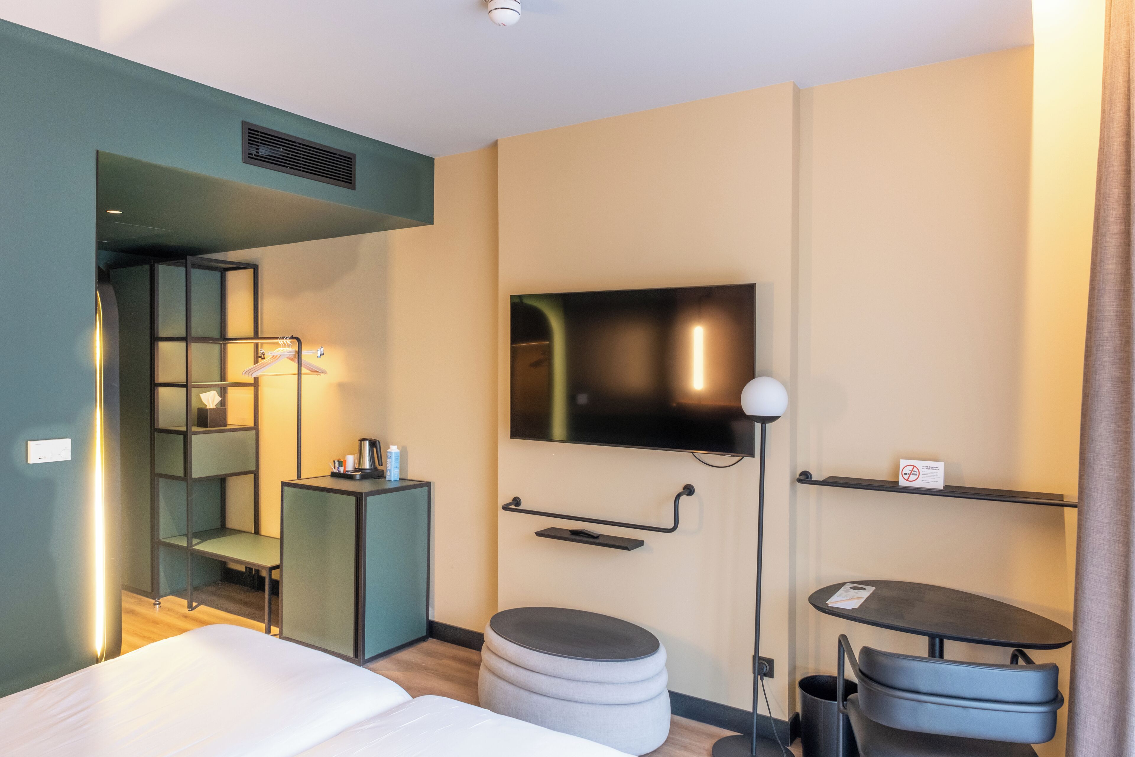 Una habitación de hotel moderna con una cama, un televisor montado en la pared, un escritorio pequeño, una silla, un estante y un otoman redondo.