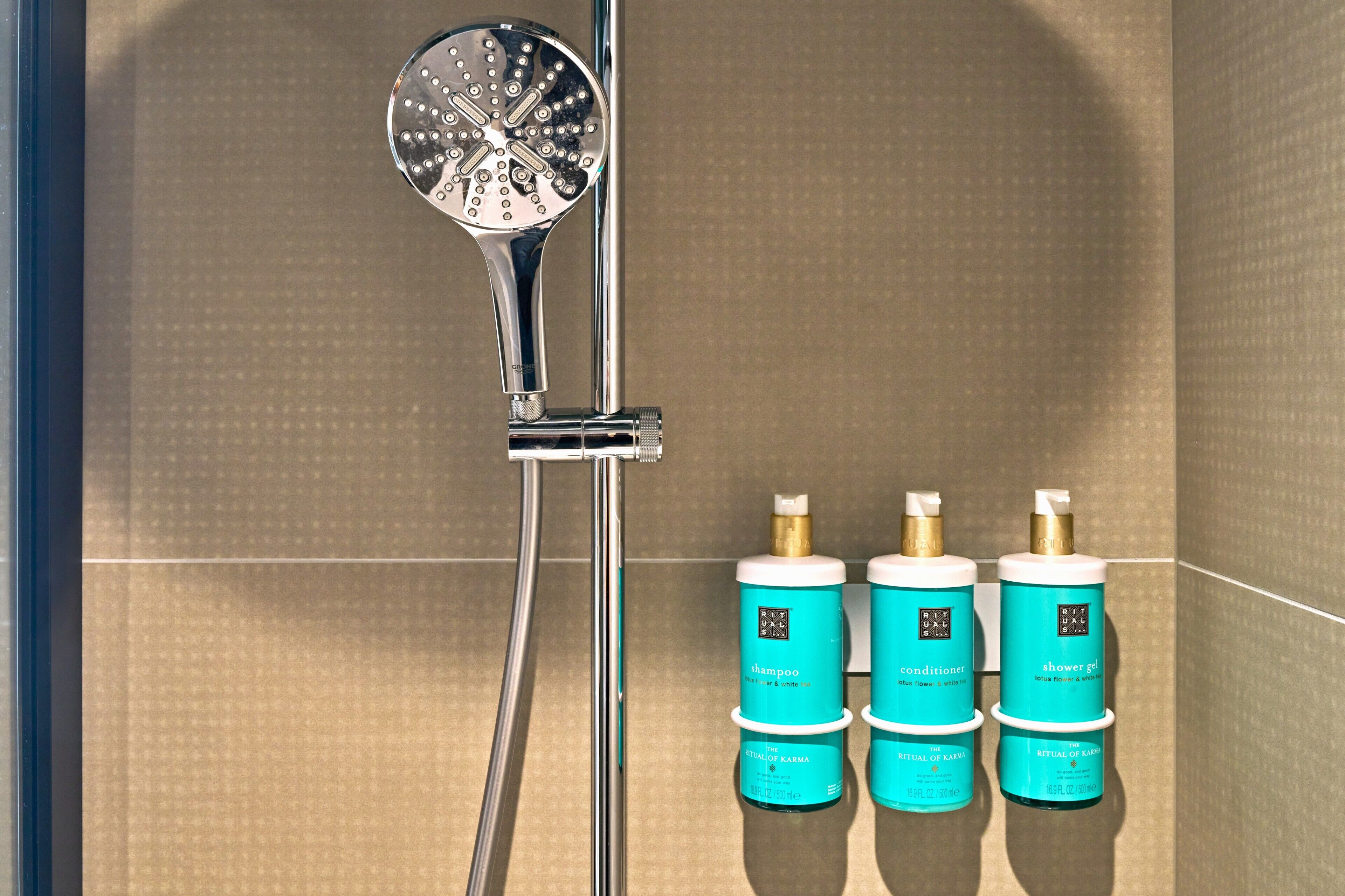 A shower with a chrome head, three bottles of Ritual of Karma products, and a tiled wall.