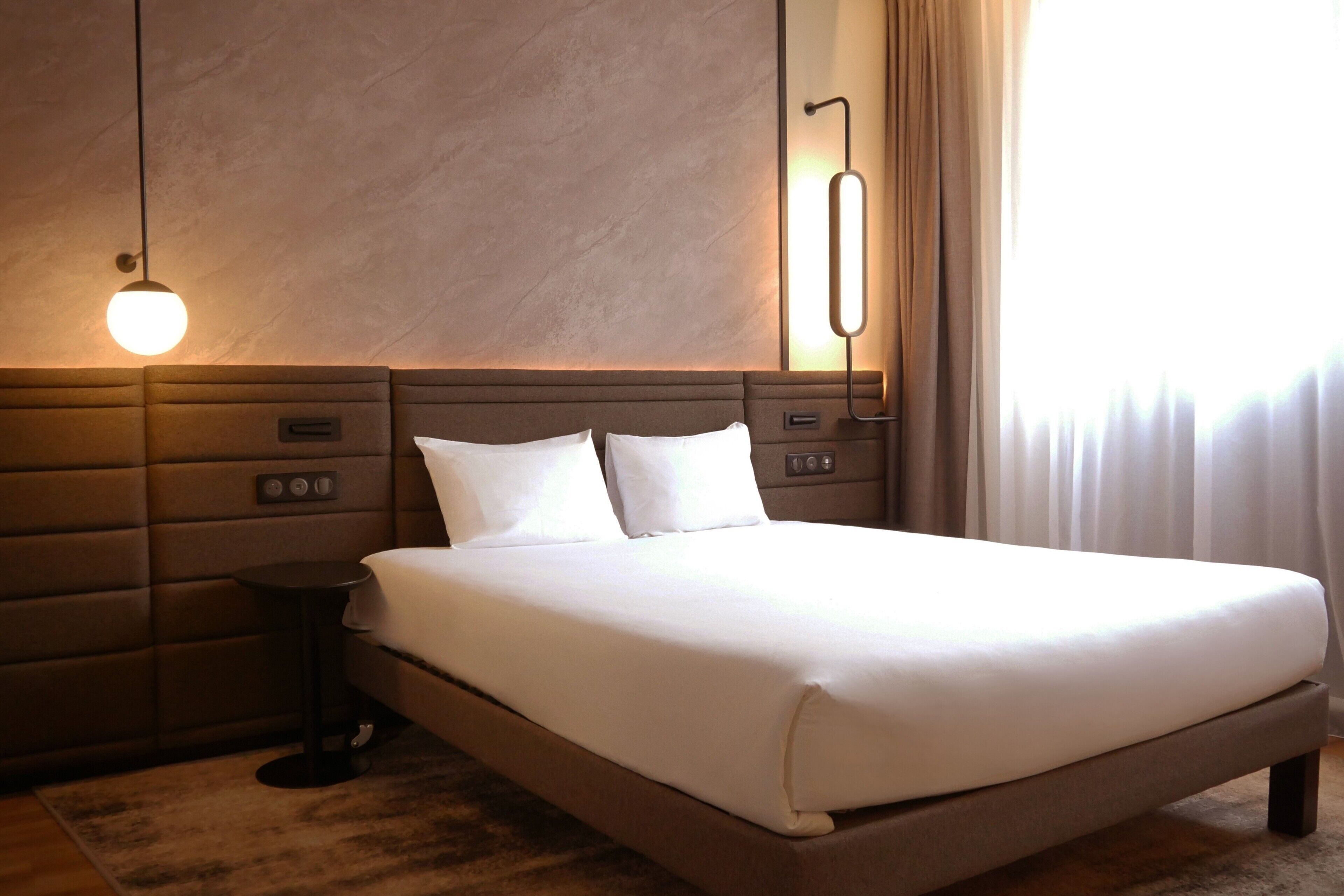 Una habitación de hotel moderna con una cama grande, una mesita de noche, una lámpara de pared y un ventanal con cortinas.