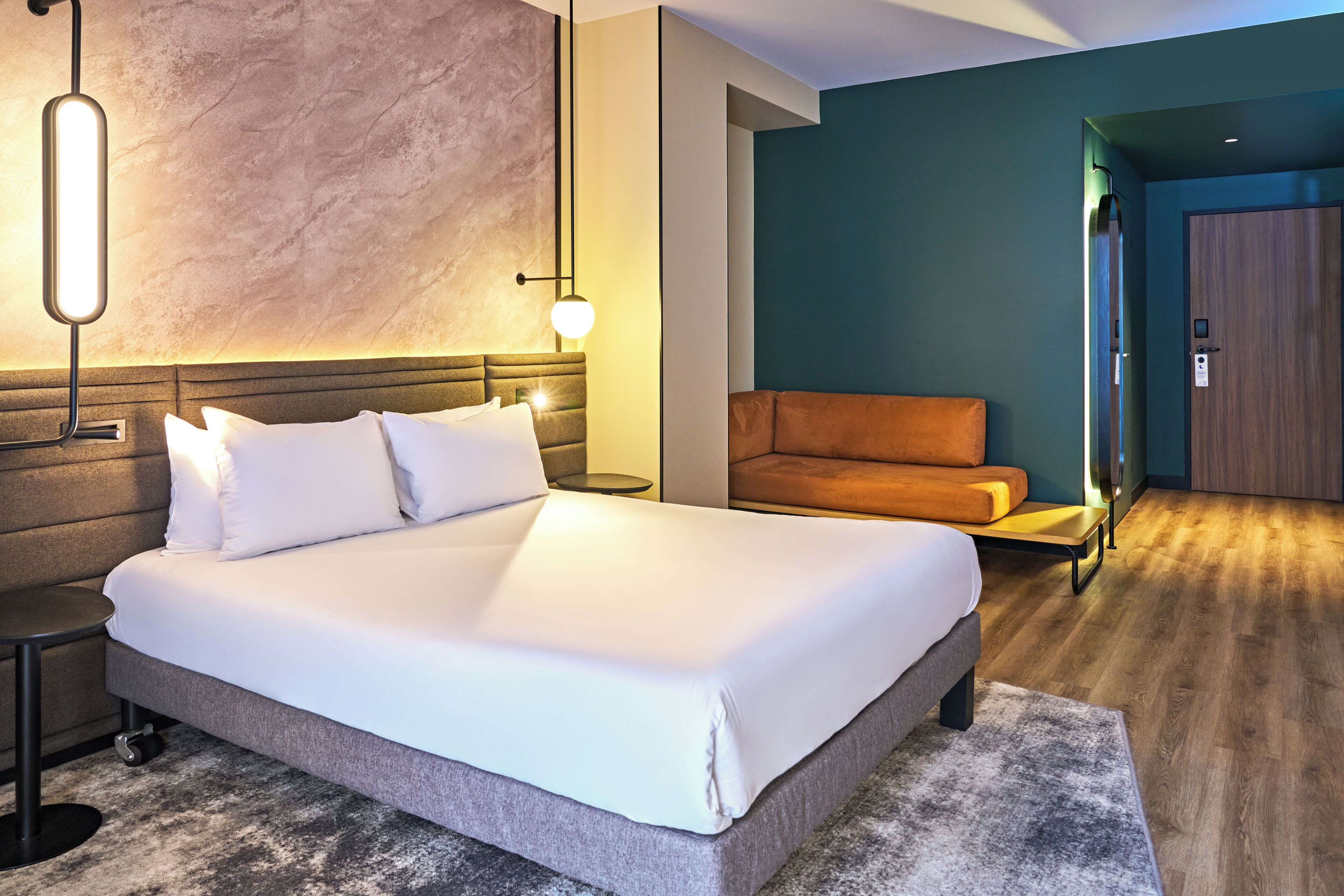 A hotel room with a large bed, a nightstand, a wall-mounted lamp, a sofa, and a wooden door.