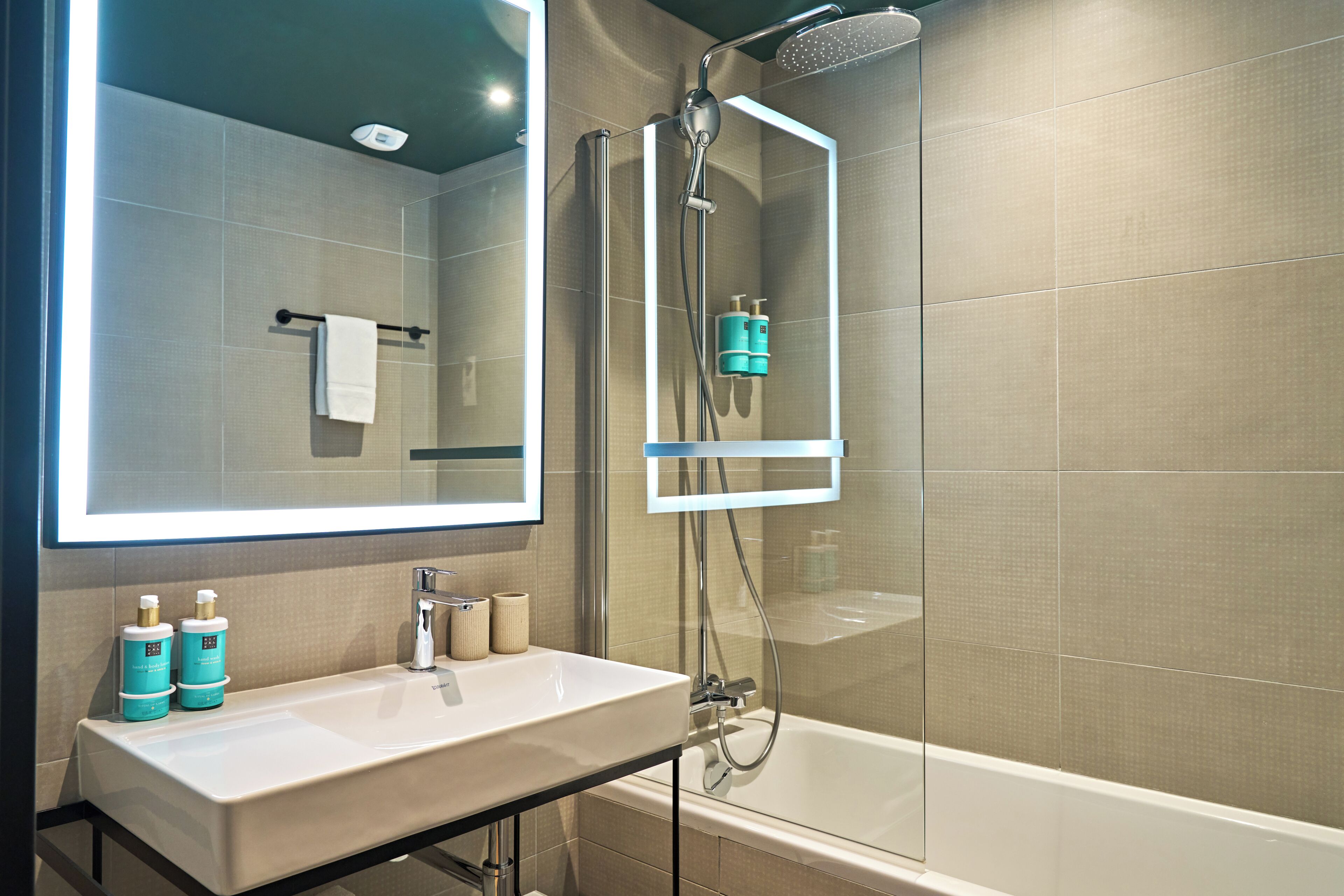 A modern bathroom with a large mirror, a sink, a shower, and two bottles of shampoo.