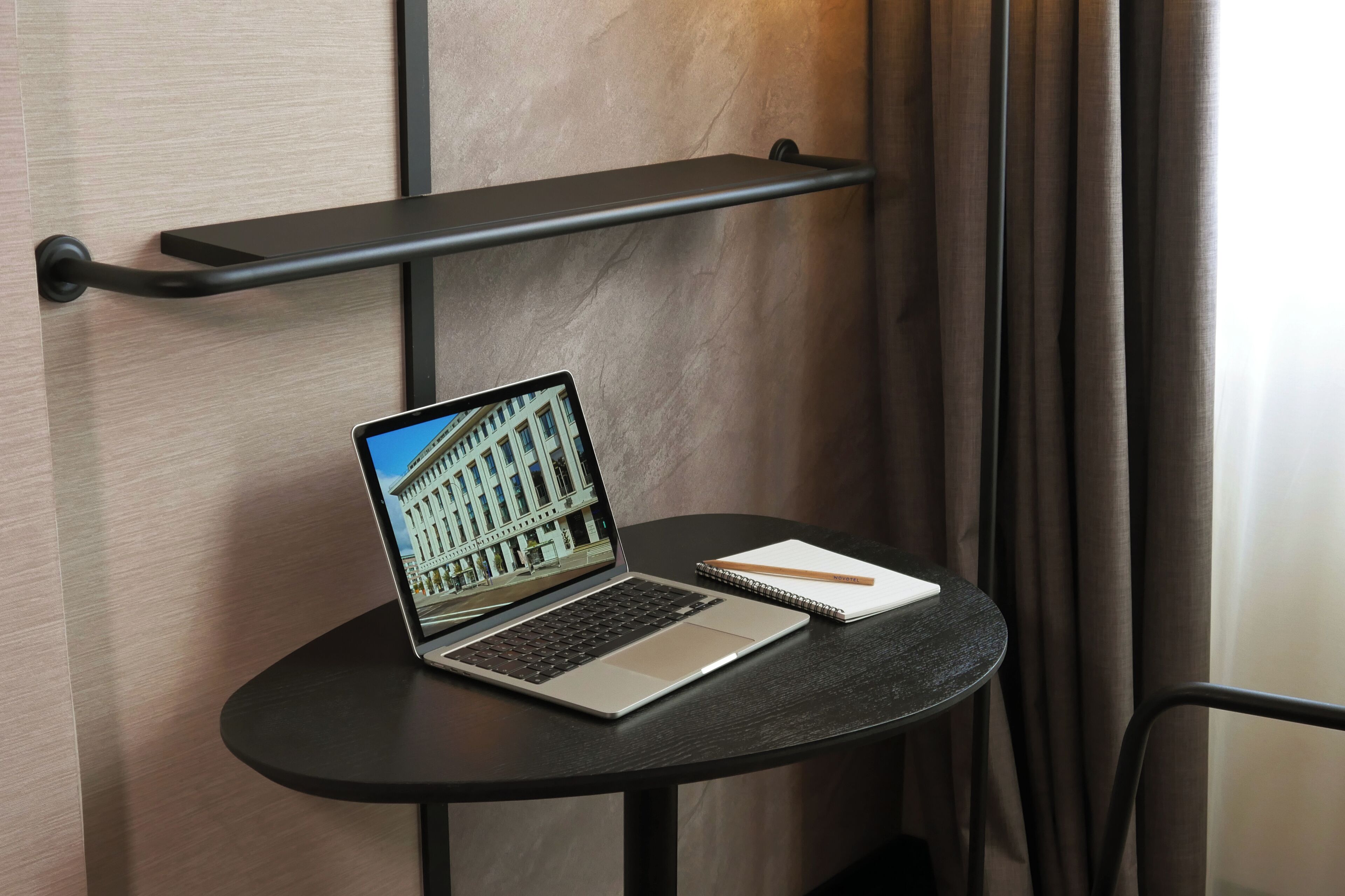 A laptop on a small table with a notebook and pencil beside it, against a wall with a shelf above.