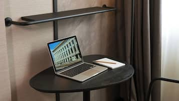 A laptop on a small table with a notebook and pencil beside it, against a wall with a shelf above.