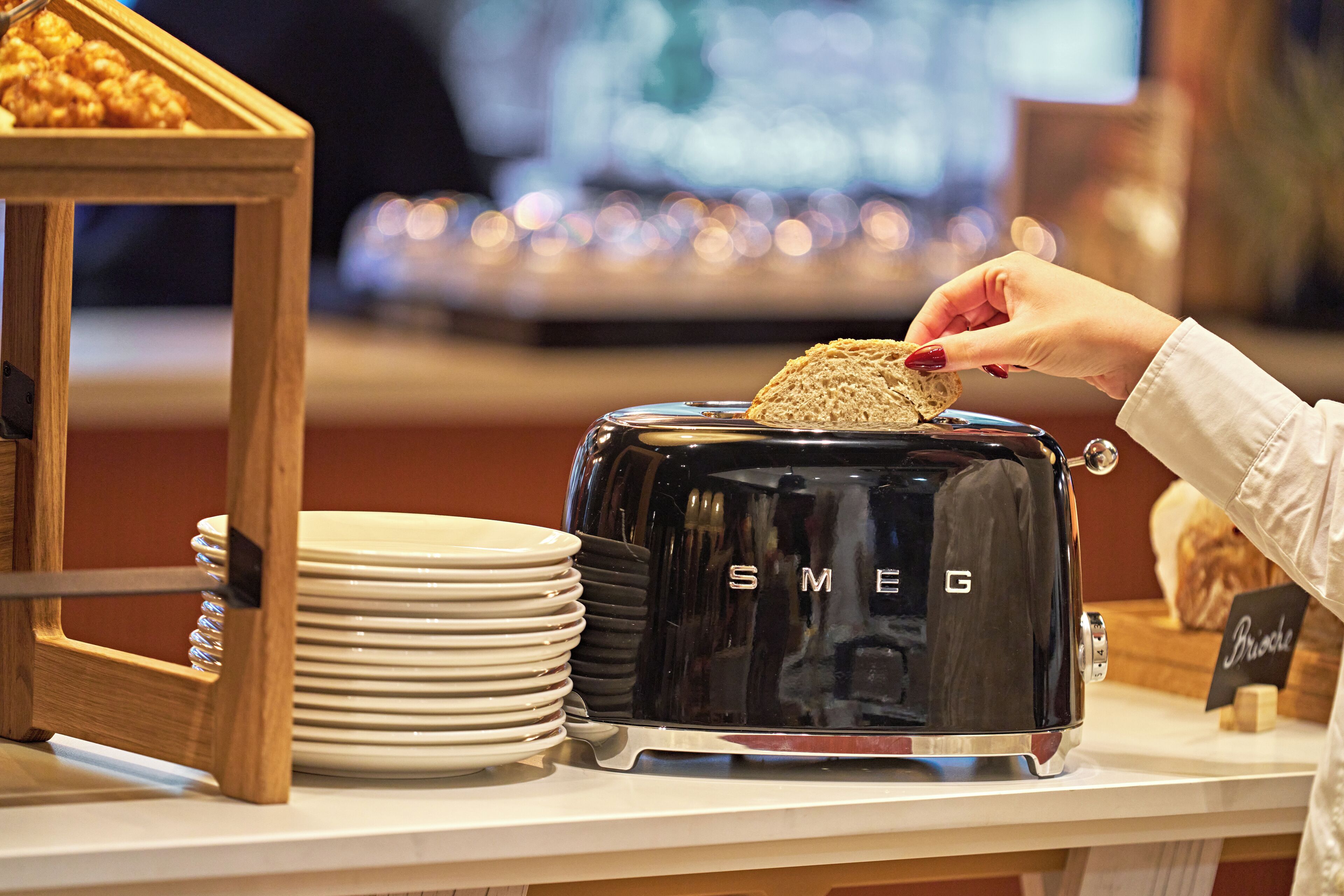 A toaster with the brand name "SMEG" on a countertop, surrounded by white plates and a wooden frame.