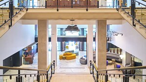 Um hall amplo com área de estar central, escada com corrimão de vidro e vista para o andar superior.
