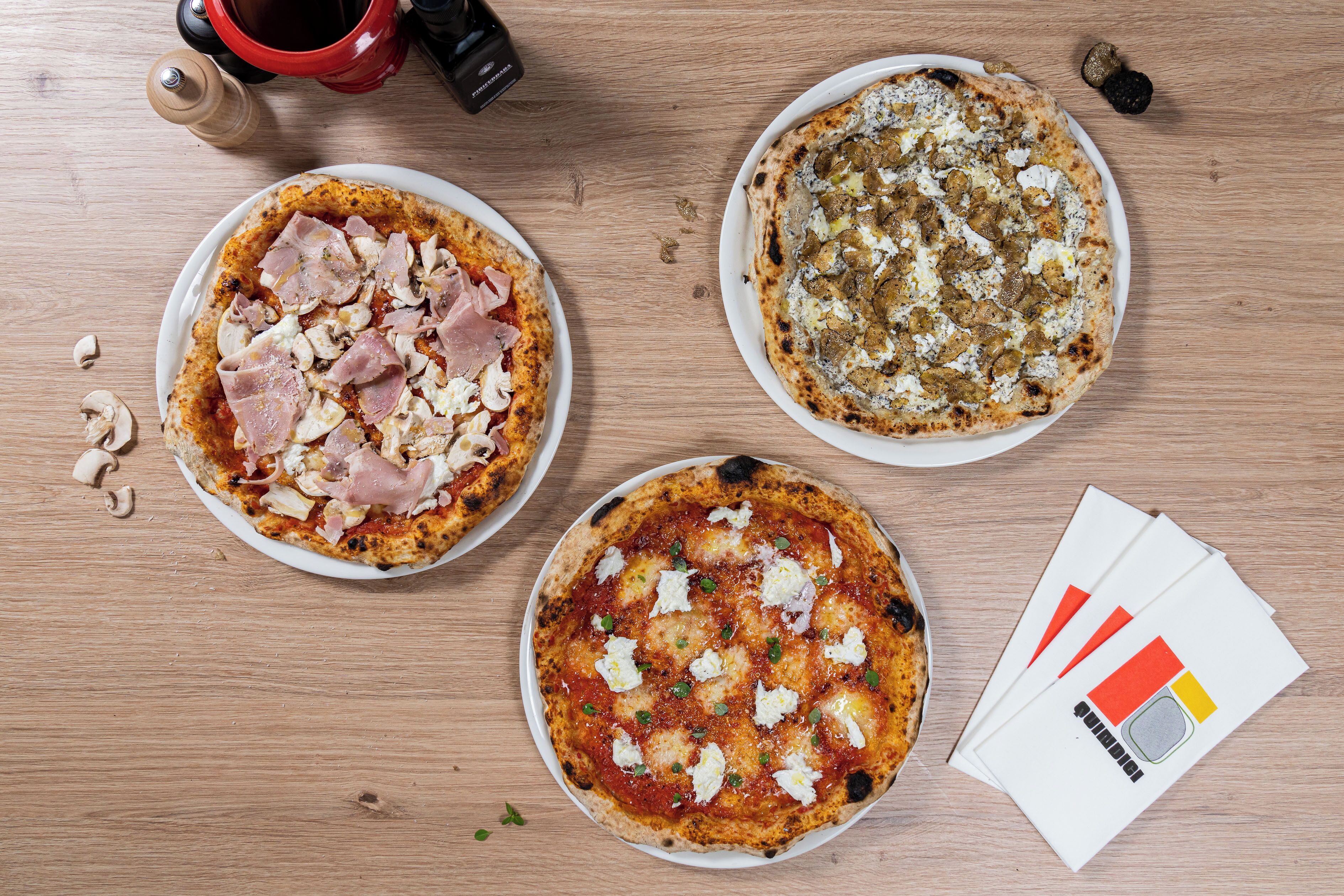 Three pizzas on plates with various toppings, a salt shaker, pepper shaker, and a bottle of olive oil on a wooden table.