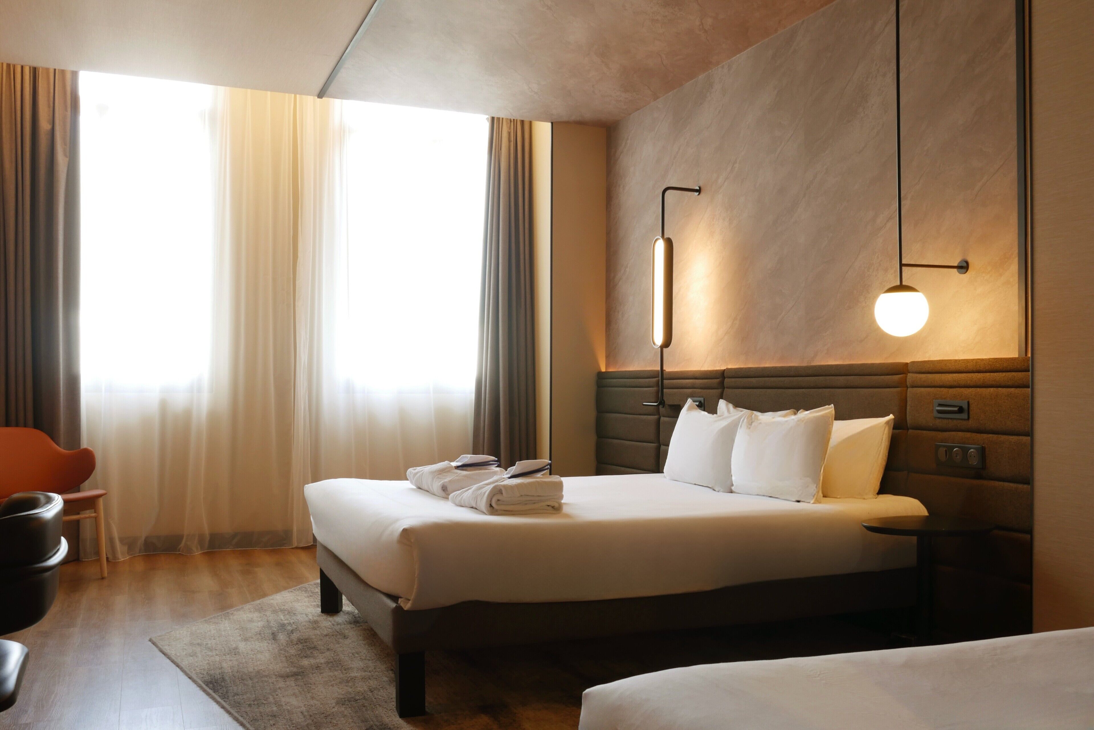 A hotel room with a bed, bedside table, lamp, curtains, and a chair.