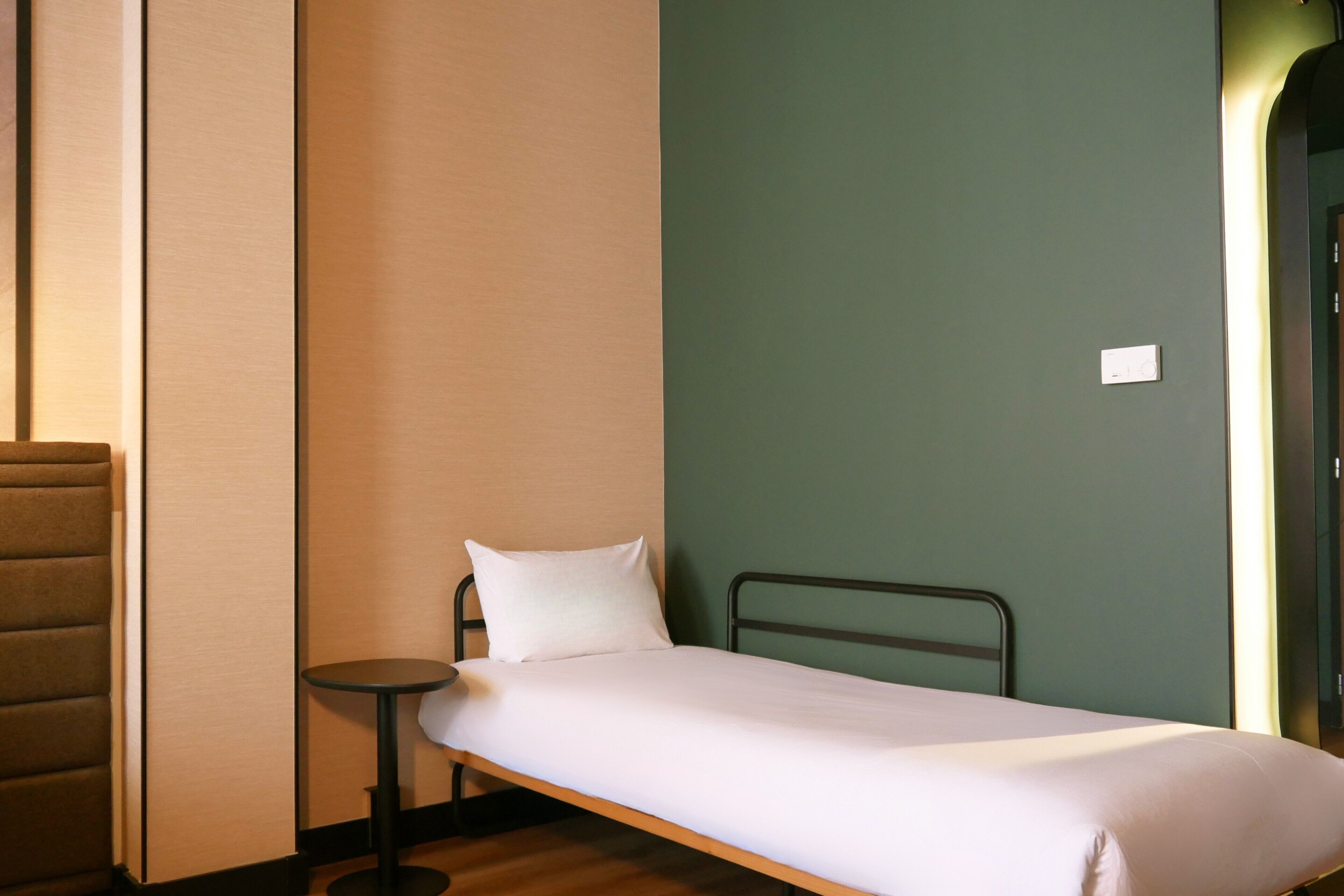A single bed with a white mattress and pillow, a small round table, and a green wall.