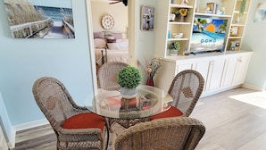 Un coin repas avec des chaises en osier et une table en verre, un lit avec vue sur la plage et un téléviseur diffusant l’émission « OCMD GETAWAYS ».