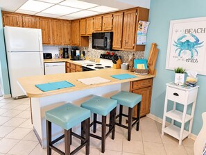 A kitchen with wooden cabinets, a white refrigerator, a microwave, a stove, and a sink. There are three bar stools with blue cushions.