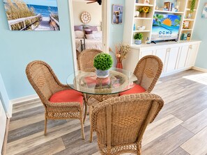 A dining area with wicker chairs and a glass table, a television on a shelf, and a view of the ocean through a window.