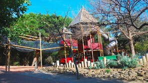 Explore the playground with rope bridges, climbing frames, and a slide set amidst a serene natural environment.